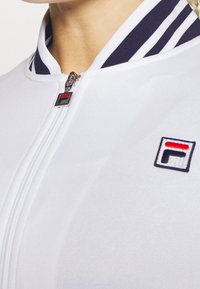 Vit bomberjacka med dragkedja och ribbade krage med marinblå ränder. Inkluderar en liten broderad logotyp på bröstet.