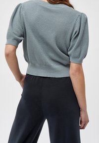 Světle modrý pletený svetr s krátkými nabíranými rukávy a žebrovaným lemem, spárovaný s tmavými kalhotami. Texturovaná tkanina je výrazná po celém kousku.