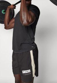 Sort atletisk skjorte med teksturerede paneler, parret med sorte shorts med en beige stribe og et hvidt PUMA-logo.