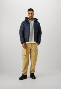 Geacă matlasată bleumarin cu glugă, combinată cu pantaloni cargo bej cu multiple buzunare și adidași negri. Ținută casual.