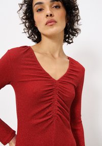 Rotes langärmliges Kleid in gerippter Textur mit tiefem V-Ausschnitt und gerafftem Detail an der Vorderseite. Der Stoff hat einen leichten Schimmereffekt.