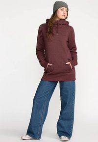 Maroon hooded sweatshirt with front pockets, paired with wide-leg blue denim jeans. Model wears a gray knit beanie. Casual outfit.