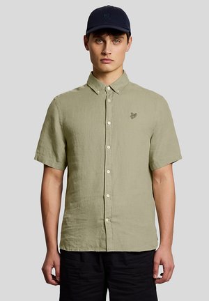 Junger Mann mit einem hellolivgrünen kurzärmeligen Hemd und einer marineblauen Kappe, der vor einem einfarbigen grauen Hintergrund steht.