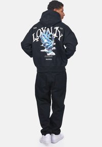 Schwarzer Hoodie mit buntem Grafikdesign eines Hand und Wolken auf dem Rücken, kombiniert mit schwarzen Hosen und weißen Sneakers.