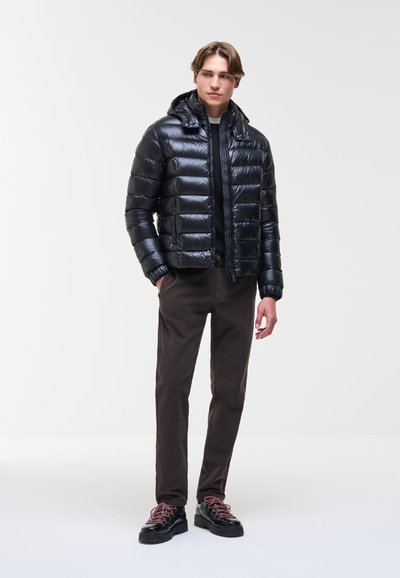Jeune homme portant une veste matelassée noire brillante, un pantalon foncé et des chaussures noires avec des lacets rouges, debout avec une main dans la poche.