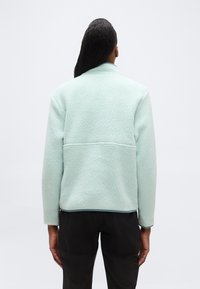 Άτομο που στέκεται με την πλάτη ορατή, φοράει ανοιχτό μπλε fleece σακάκι με υφή και μαύρο παντελόνι, σε απλή ανοιχτή φωτεινή φόντο.