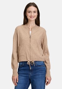 Beige Strickjacke mit Reißverschluss, runder Ausschnitt, zwei Fronttaschen und Kordelzug am Saum. Weicher Stoff und entspannter Schnitt; lange Ärmel.