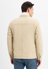 Beige, leichtgewichtige Jacke mit Kragen, langen Ärmeln und matter Oberfläche, die eine sichtbare Naht auf dem Rücken und ein neutrales Design aufweist.