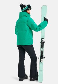Green insulated jacket, black pants, holding mint-colored skis with black bindings and patterned beanie. Snow goggles resting on forehead.