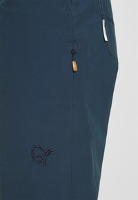 Navy blue outdoor pants with a drawstring detail and wooden toggle. Features an embroidered logo in a contrasting color near the pocket.