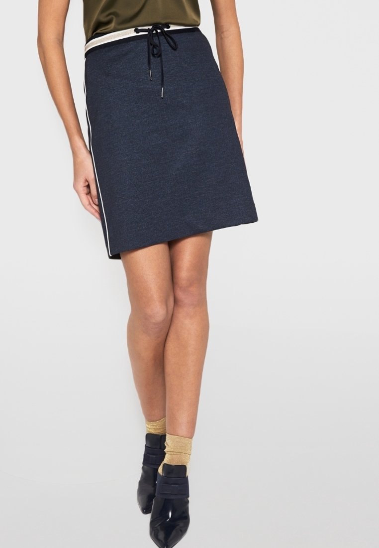 Laivastonsininen A-mallinen hame, jossa on teksturoitu pinta, nyörivyötärö ja valkoiset sivuraidat. Yhdistetty kiiltäviin mustiin kenkiin ja kultaisiin sukkiin.