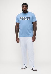 Tricou din bumbac de un albastru deschis, cu "POLO" imprimat în litere îndrăznețe de culoare bleumarin, asortat cu pantaloni albi cu dungi și adidași albi cu accente negre.
