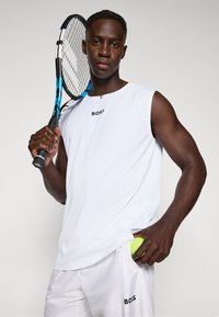 Λευκή αμάνικη αθλητική μπλούζα με το λογότυπο "BOSS", σε συνδυασμό με λευκές βερμούδες. Το μοντέλο κρατά μια μπλε ρακέτα τένις και μια κίτρινη μπάλα τένις.