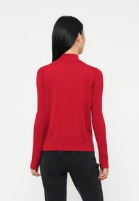 Frau mit langen schwarzen Haaren, die einen roten Langarmpullover und schwarze Jeans trägt, von hinten vor einem schlichten Hintergrund gezeigt.