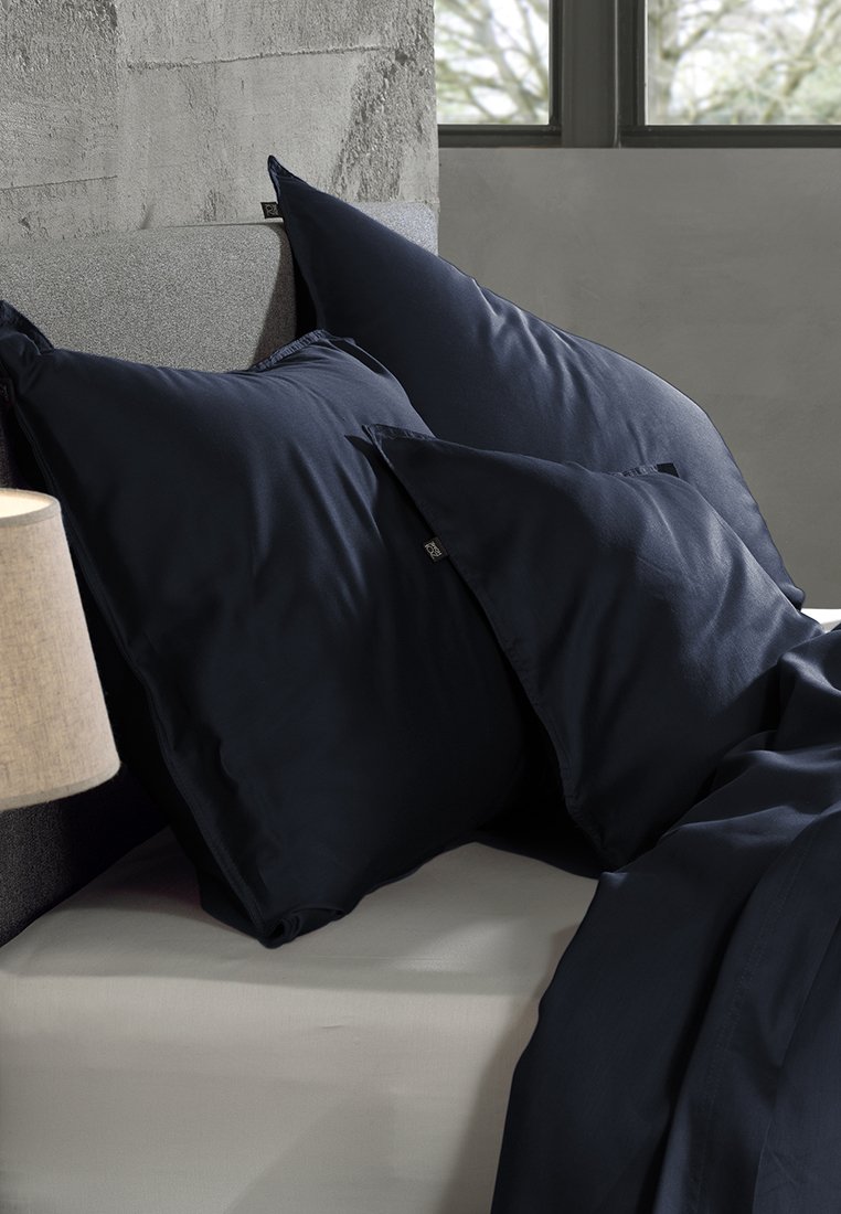 Tre cuscini blu scuro disposti su un letto grigio chiaro, con una lampada beige e una finestra sullo sfondo.