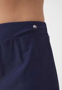 Kovinska modra elastika iz gladkega materiala, ki ima majhen, okrogel kovinski logotip na hrbtni strani. Material je elastičen in gladek.