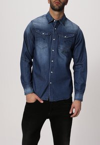 Jeansskjorta med en mörkblå tvätt, långa ärmar, spetsig krage, två bröstfickor och knäppning fram.