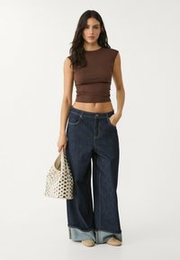 Brun tajt topp, vida mörka jeans med ljusa denimärmmar, och en beige väska med silvernitar. Modell som håller väskan, stående mot en enkel bakgrund.