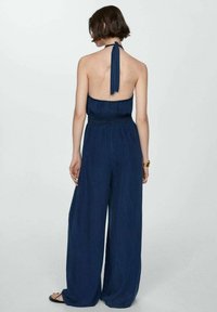 Námořnická modrá overal bez rukávů s otevřeným zadním dílem, nařaseným pasem a širokými nohavicemi. Hladká látka s lehkým textura.