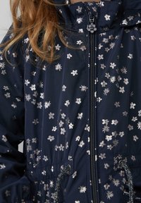 Granatowa kurtka z srebrnym wzorem kwiatowym, z zamkiem błyskawicznym z przodu, z regulowanym pasem i kapturem. Gładki materiał o lekko połyskującej powierzchni.