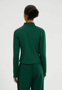 Top verde de manga larga con cuello alto, textura suave y un dobladillo ligeramente curvado en la parte posterior. La tela parece ligera y flexible.