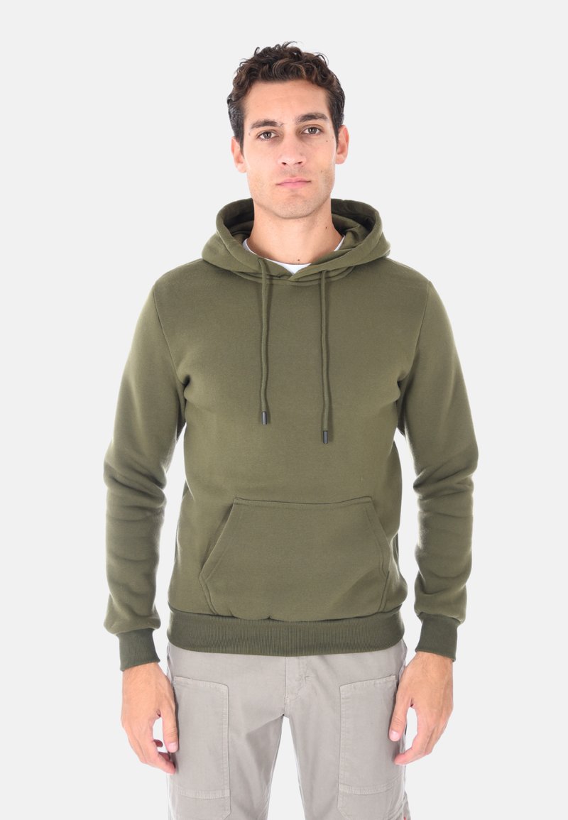 Uomo con felpa verde oliva e pantaloni cargo grigi chiari, in piedi e rivolto in avanti con espressione neutra su sfondo semplice.