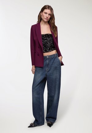 Blazer viola su misura sopra un top corto nero con paillettes, abbinato a jeans blu in denim dal taglio lungo e scarpe nere con punta e decorazioni.