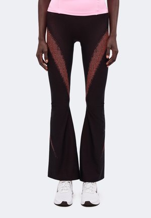 Personne portant un pantalon évasé noir avec des motifs chevrons roses en pointillés et des baskets blanches, debout sur un fond clair uni.