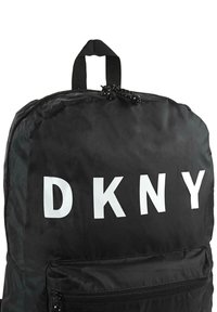 Mochila de nailon negra con letras grandes en blanco "DKNY", un asa superior y un bolsillo frontal con cremallera negra.