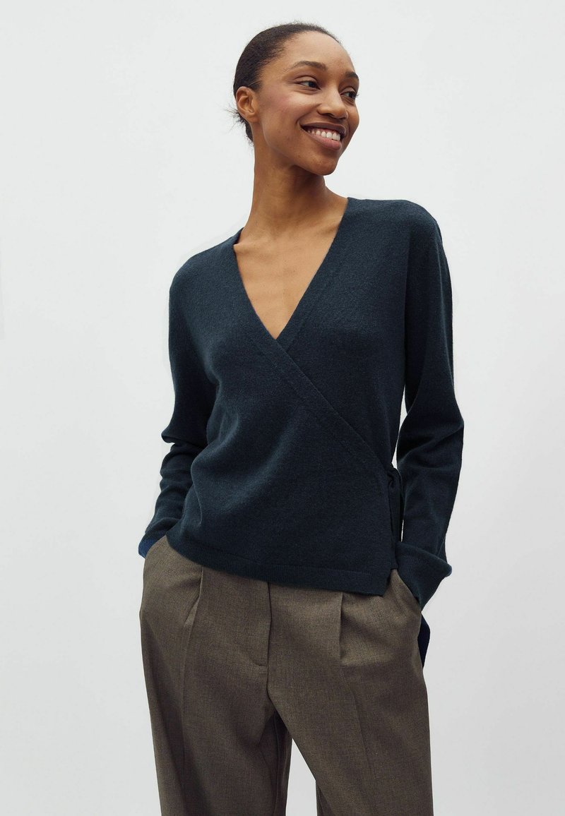 Smejoša sieviete ar rokām kabatās, valkājot tumši zilas apkakles džemperi un brūnas vaļīgas bikses uz vienkārša gaiša fona.