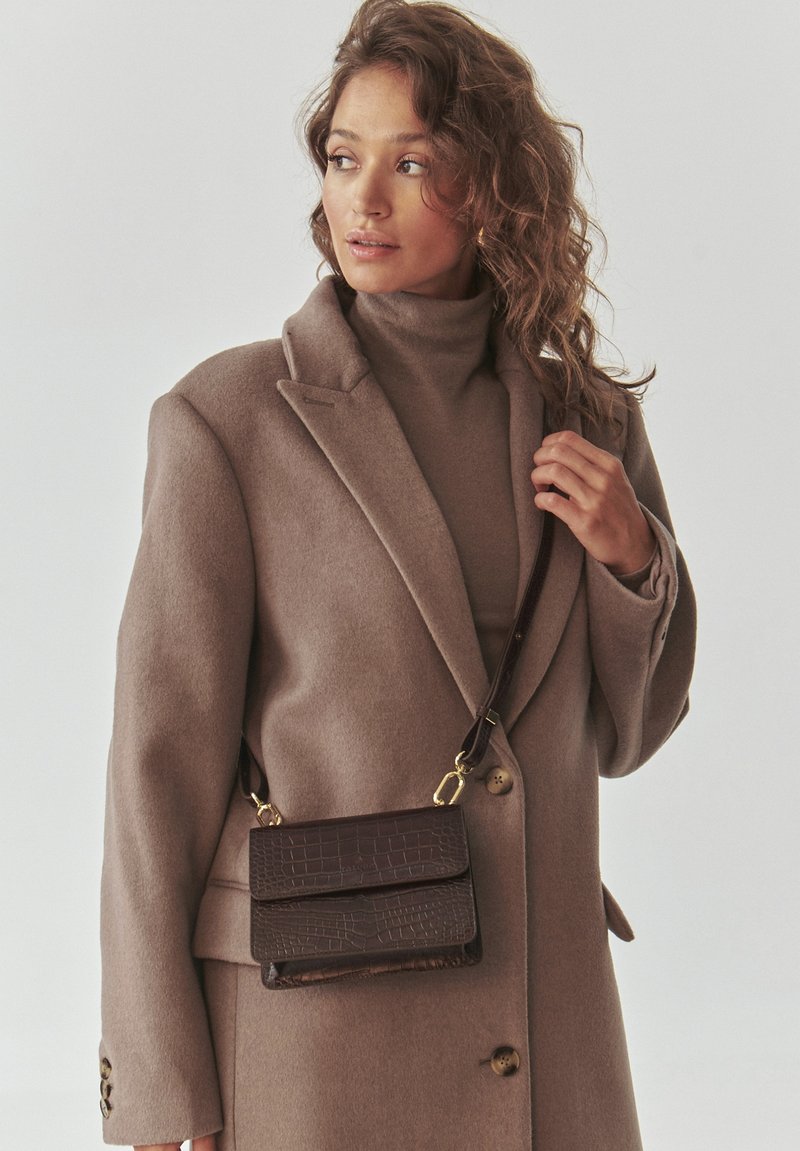 Borsa a tracolla in pelle con stampa a coccodrillo marrone, con dettagli in oro, forma rettangolare e due scomparti, indossata da un modello con un cappotto beige.