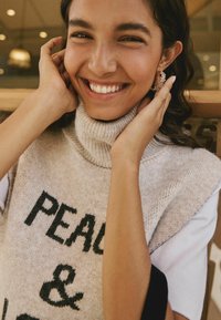 Maglione in beige con testo nero "PEACE &", con colletto alto, abbinato a orecchini a cerchio dorati scintillanti. Persona sorridente mentre si tocca il viso.