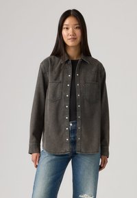 Νεαρή γυναίκα με μακριά σκουρόχρωμα μαλλιά, ντυμένη με μαύρο τζιν πουκάμισο με κουμπιά πάνω από μαύρο τοπ και μπλε σκισμένο τζιν, στεκόμενη μπροστά σε απλό φόντο.