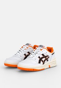 Baskets blanches de sport avec des accents noirs et orange, boîte à orteils perforée, semelle en caoutchouc texturée, design à lacets et logo sur la languette.