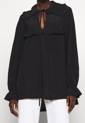 Schwarze langärmlige Bluse mit gerafftem Kragen und Manschetten, plissiertem Frontdetail, gebundenem Ausschnitt, getragen mit weißen Hosen von einer Person mit dunkler Hautfarbe.