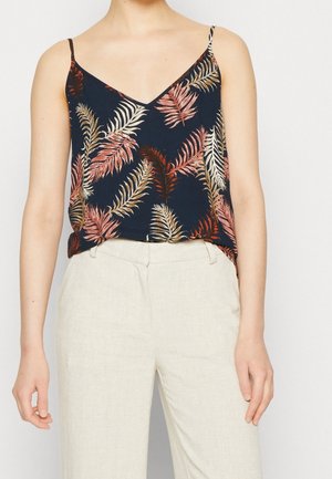 Persoon die een marineblauwe mouwloze top met roze en beige bladpatronen draagt, ingestopt in een hooggetailleerde off-white broek, staand tegen een effen achtergrond.