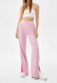 Pink, high-waisted trousers with a straight cut, featuring side slits and a smooth texture, paired with white sneakers and a cropped top.