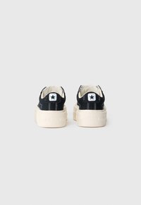 Black canvas sneakers with white rubber soles, featuring a star logo on the heel and stitching accents. Low profile and minimalist design.