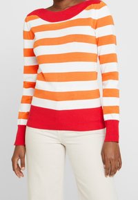 Pull rayé avec des rayures horizontales orange et blanches, un col et des poignets côtelés rouges, et une coupe ajustée.