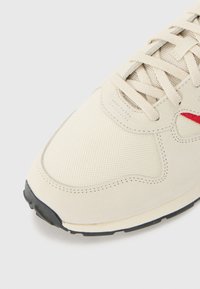 Beige sneakers med en blanding af glat ruskind og tekstureret mesh. Rød detalje nær snøringerne. Sort gummisål med lyse beige detaljer.