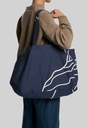 Persoon die een bruine trui en blauwe spijkerbroek draagt met een grote marineblauwe draagtas met witte abstracte lijnpatronen op de schouder.