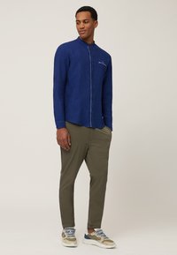 Marineblauw linnen overhemd met een bandkraag, sluiting met knopen aan de voorkant en witte stiksels. Gepaard met olijfgroene broeken en sneakers.