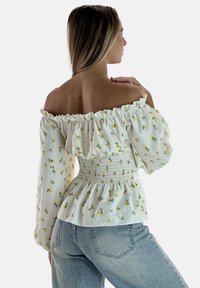 Weiße Schulterfreie Bluse mit Blumenmuster, gerafftem Oberteil und aufgeplusterten Ärmeln. Kombiniert mit hellen Jeans aus Denim.