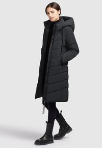 Sort dunjakke med hætte, med en lang design, kviltet tekstur, sidelommer og lynlås. Båret med sorte ankelstøvler.