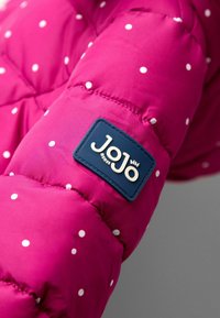 Fuchsia Pufferjacke mit Ärmel und weißen Punkte, versehen mit einem blauen Etikett mit "Jojo" in cremefarbener Schrift. Weicher, gestepptes Material.