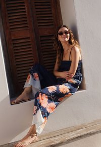 Salopetă florală bleumarin cu flori supradimensionate în nuanțe de roz și portocaliu. Are un corset cu volane, picioare largi și accente transparente la tiv.