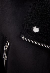 Svart jacka med slät tyg och en texturerad fleecekrage, silverdragkedjedetalj och en liten silveraccent vid sömmen.