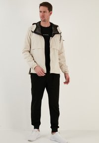 Lichtbeige windbreaker met een rits aan de voorkant, elastische manchetten en twee zakken aan de voorkant, gedragen over een zwart t-shirt en gecombineerd met zwarte joggers en witte sneakers.