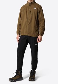 Bruine softshell jas met hoge kraag, ritszak op de borst; zwarte waterdichte broek; robuuste bruine wandelschoenen met veter en profiel.