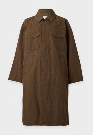 Un mannequin masculin pose devant un fond de studio uni, vêtu d’un long manteau marron, d’un pull bleu marine à torsades, d’un pantalon crème, d’une casquette foncée et de mocassins noirs.